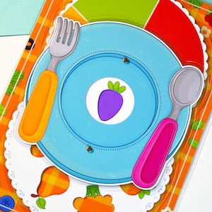 May include: A colorful children's educational toy featuring a blue plate with a purple carrot graphic, a green, red, and yellow food guide, and a fork and spoon with orange and pink handles. The background has an orange plaid pattern.