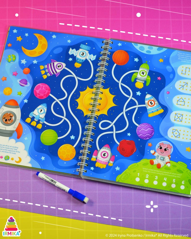 May include: A spiral-bound activity book with a space theme, featuring a maze with rockets, planets, and a sun. A blue and white marker lies on the purple surface. The book's pages are filled with colorful illustrations and the text "IRMIKA".