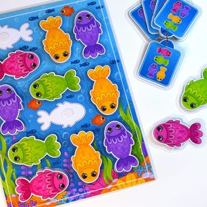 Puede incluir: Un rompecabezas infantil con peces de dibujos animados en varios colores, incluyendo naranja, morado, verde y rosa. El rompecabezas tiene un fondo azul con temática submarina e incluye tarjetas a juego.