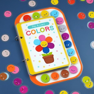 May include: A colorful children's learning set featuring a spiral-bound book titled "Colors" with a flower illustration. The set includes a tray with smiling face stickers in various colors, perfect for educational play.