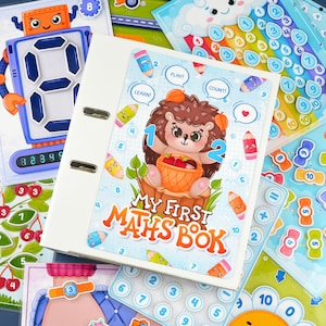 Puede incluir: Una carpeta blanca con el texto "MY FIRST MATHS BOOK" en la portada, con un erizo de dibujos animados sosteniendo una cesta de fresas. Tarjetas coloridas con números y matemáticas rodean la carpeta.