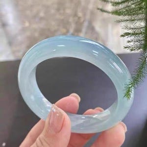 May include: A pale blue jade bangle. The translucent bracelet is held in a hand, highlighting its polished surface. The jade has a subtle, cool colour, and the bracelet's circular shape is clearly visible.