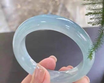 May include: A light blue jade bangle bracelet. The smooth, translucent bracelet is held in a hand, showcasing its polished surface. The jade has a subtle, cool color, and the bracelet's circular shape is clearly visible.