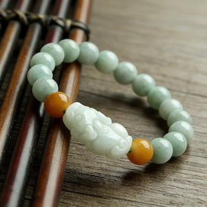 May include: A jade bracelet featuring pale green beads and two orange beads flanking a carved white jade figurine. The bracelet is displayed on a wooden surface, showcasing the natural colors and textures of the materials.