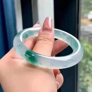 May include: A jade bangle bracelet, displaying a blend of light green and white tones. The bracelet is held in a hand, highlighting its smooth, polished finish. The circular bracelet is made of jade and has a translucent appearance.