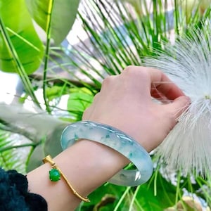 May include: A hand adorned with a gold bracelet featuring a green jade charm and a large, translucent jade bangle. The jade displays a pale green and blue colour. The background includes green foliage and white feathery accents.