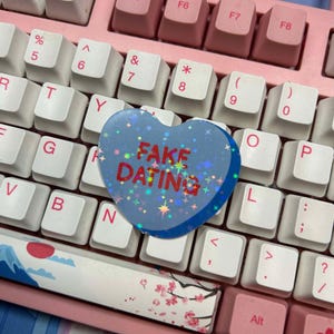 Puede incluir: Un caramelo azul en forma de coraz&oacute;n con un acabado hologr&aacute;fico y el texto "FAKE DATING" en letras rojas. El caramelo est&aacute; sobre un teclado rosa y blanco.