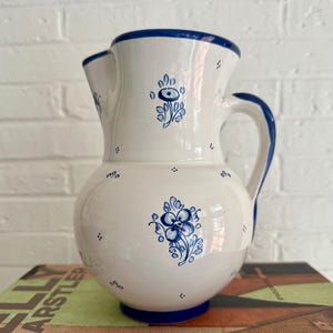 Może przedstawiać: Biały ceramiczny dzban z niebieskimi wzorami kwiatowymi i niebieskim uchwytem. Dzban ma zaokrąglony korpus i dzióbek. Obręcz i podstawa są również niebieskie. Dzban jest ozdobiony niebieskimi wzorami kwiatowymi i małymi niebieskimi kropkami.