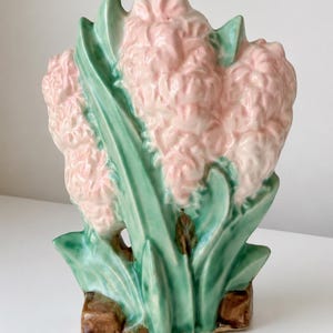 May include: A ceramic figurine shaped like a cluster of three pale pink hyacinth flowers with long green leaves and stems, set upon a brown base. The piece features a glossy finish and molded details that define the petals and foliage texture.