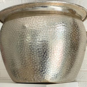 May include: A large, silver metal ice bucket with a hammered texture. The bucket has a wide, rounded shape with a flared rim and two lion head handles with ring pulls. The bucket is sitting on a white surface.