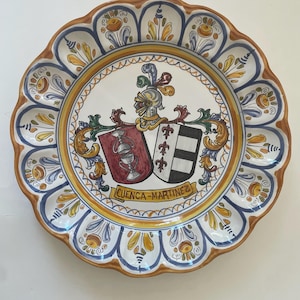 May include: A decorative ceramic plate with a scalloped edge. The plate features a central crest design with a helmet, shields, and the text "CUENCA-MARTINEZ". The rim is decorated with blue, yellow, and brown floral patterns.