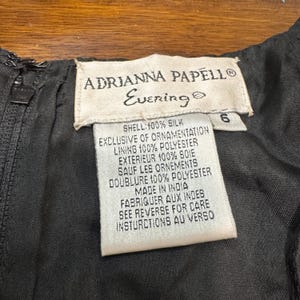 May include: Close-up of a black evening dress label. The label reads "ADRIANNA PAPELL Evening" and includes fabric composition details: shell 100% silk, lining 100% polyester. The label also states "Made in India".