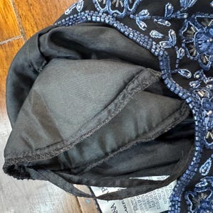 May include: Close-up of a black garment with blue beaded floral embroidery. The lining is black. The garment has a drawstring. The tag reads "NA" and "UER".