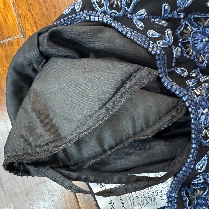 May include: Close-up of a black garment with blue beaded floral embroidery. The lining is black. The garment has a drawstring. The tag reads "NA" and "UER".