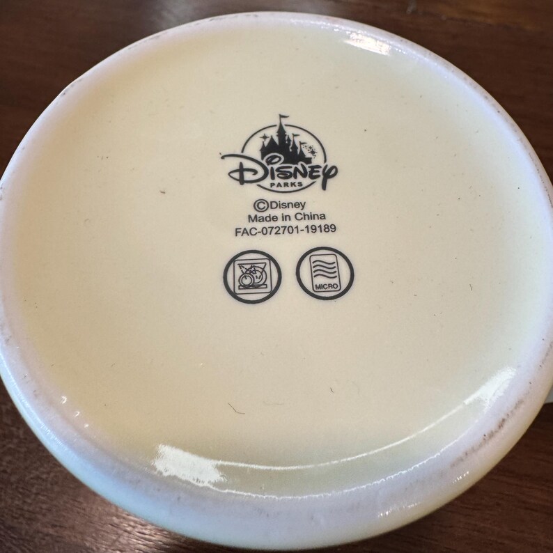 Op de afbeelding: De onderkant van een cr&egrave;mekleurige mok met het Disney Parks-logo, een kasteelsilhouet en de tekst "Disney". Onder het logo staat "Made in China" en "FAC-072701-19189". Er zijn twee symbolen, &eacute;&eacute;n voor vaatwasmachinebestendig en &eacute;&eacute;n voor magnetronbestendig.