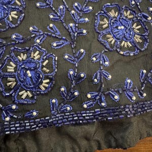 May include: Close-up of a black fabric with intricate blue beaded floral and leaf designs. The beads are a deep blue with silver accents, creating a textured and detailed pattern. The bottom edge has a row of blue beads.