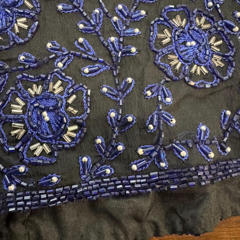 May include: Close-up of a black fabric with intricate blue beaded floral and leaf designs. The beads are a deep blue with silver accents, creating a textured and detailed pattern. The bottom edge has a row of blue beads.