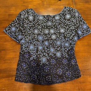 May include: Black short-sleeved top with intricate blue floral embroidery. The design features a pattern of flowers and leaves, with a gradient effect from light to dark blue. The top has a round neckline and is made of a lightweight fabric.