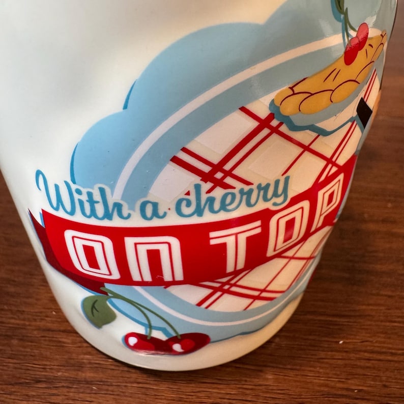 Op de afbeelding: Witte keramische mok met een retro-ontwerp. De mok heeft een rode banner met de woorden "ON TOP" in witte blokletters en de zin "With a cherry" in een scriptlettertype. Een cartoon kersentaart en kersen zijn ook zichtbaar.