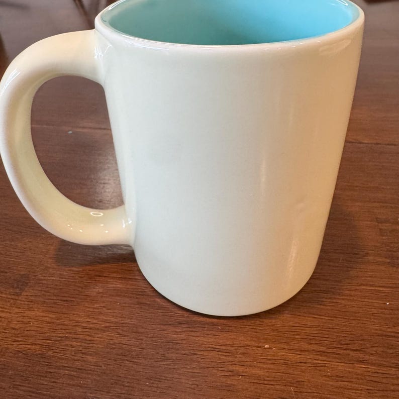 Op de afbeelding: Een lichtgroene keramische mok met een helderblauwe binnenkant. De mok heeft een groot, gebogen oor en een gladde, glanzende afwerking. Ontworpen voor warme dranken.