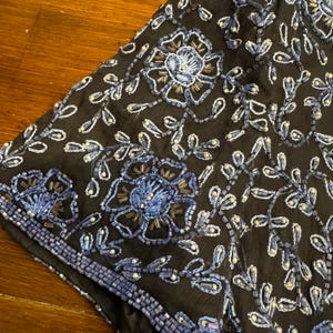 May include: Close-up of a black fabric with intricate floral embroidery. The design features large blue flowers and delicate white and blue beaded accents. The edges are trimmed with a beaded border, creating a detailed and elegant look.
