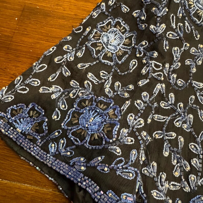 May include: Close-up of a black fabric with intricate floral embroidery. The design features large blue flowers and delicate white and blue beaded accents. The edges are trimmed with a beaded border, creating a detailed and elegant look.