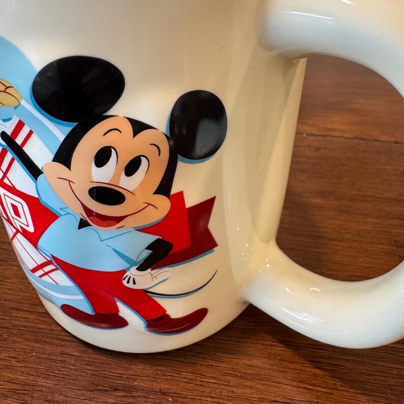 Op de afbeelding: Cr&egrave;mekleurige keramische mok met een lachende Mickey Mouse-afbeelding. Mickey draagt een blauw overhemd, een rode broek en een rode cape. De mok heeft een grote, gebogen handgreep en een glanzende afwerking.