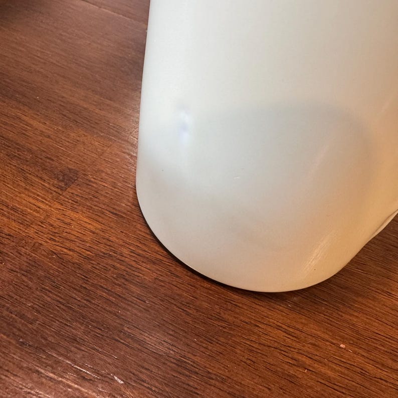 Op de afbeelding: Een gebogen, cr&egrave;mekleurig keramisch object met een glad oppervlak. Het object staat tegen een achtergrond van een donkerbruin houten oppervlak. De afgeronde vorm suggereert een deel van een groter vat, zoals een vaas of een mok.