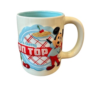 Op de afbeelding: Cr&egrave;me keramische mok met een lichtblauwe binnenkant en een grote handgreep. De mok heeft Mickey Mouse en de tekst "With a cherry ON TOP" in rood en blauw. Een kersentaartafbeelding is ook inbegrepen.