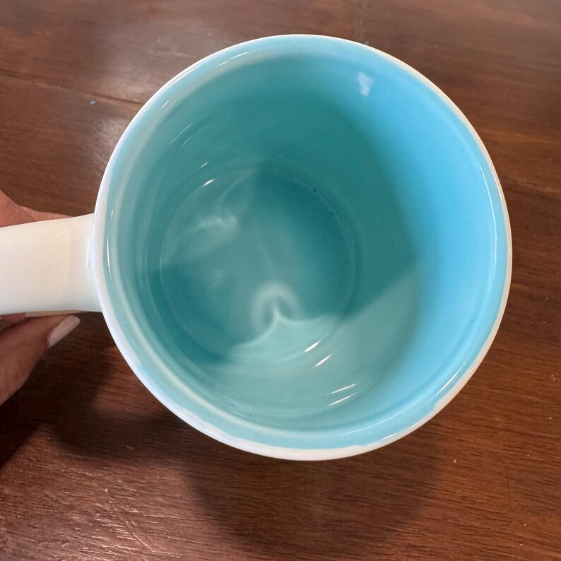 Op de afbeelding: Een witte keramische mok met een lichtblauwe binnenkant. De mok heeft een wit handvat en een gladde, glanzende afwerking. De mok is leeg en lijkt schoon te zijn.