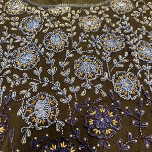 May include: Close-up of a black fabric with intricate floral embroidery. The design features blue and gold beaded flowers and leaves, creating a detailed and textured pattern. The embroidery is densely packed, covering the surface with a rich, decorative effect.