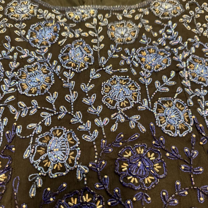 May include: Close-up of a black fabric with intricate floral embroidery. The design features blue and gold beaded flowers and leaves, creating a detailed and textured pattern. The embroidery is densely packed, covering the surface with a rich, decorative effect.