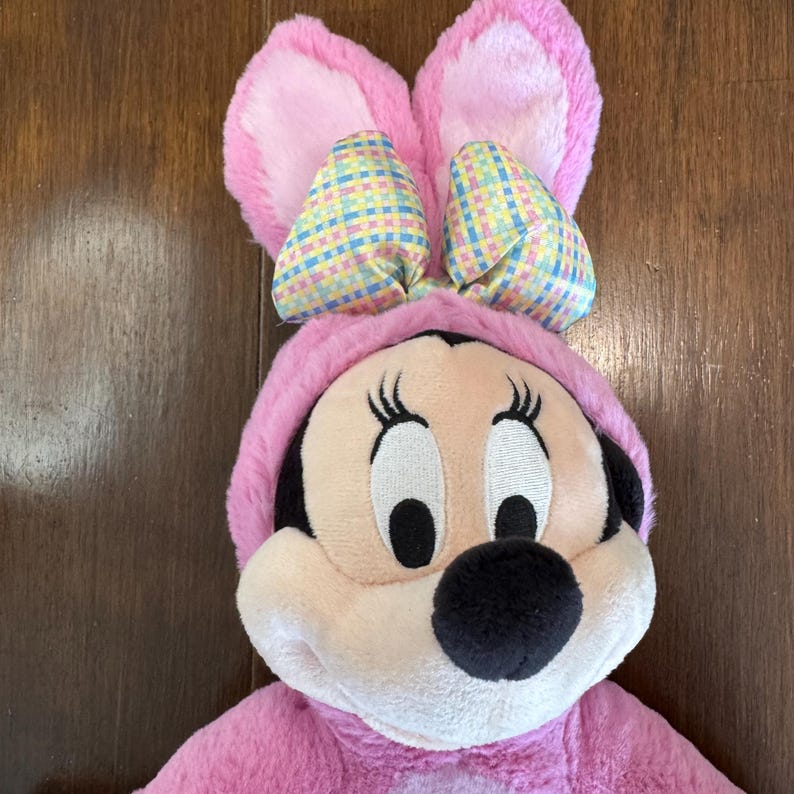 May include: A plush Minnie Mouse dressed in a pink bunny costume. The costume features large pink bunny ears with pastel plaid bows. Minnie's face is visible, with her signature black nose, eyelashes, and rosy cheeks. The background is a wooden surface.