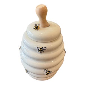 Può includere: Un vasetto per miele in ceramica color crema a forma di alveare con finitura lucida. Il vasetto presenta piccole api dipinte a mano in nero e giallo sparse sull'esterno a coste e include un manico in legno chiaro sul coperchio.