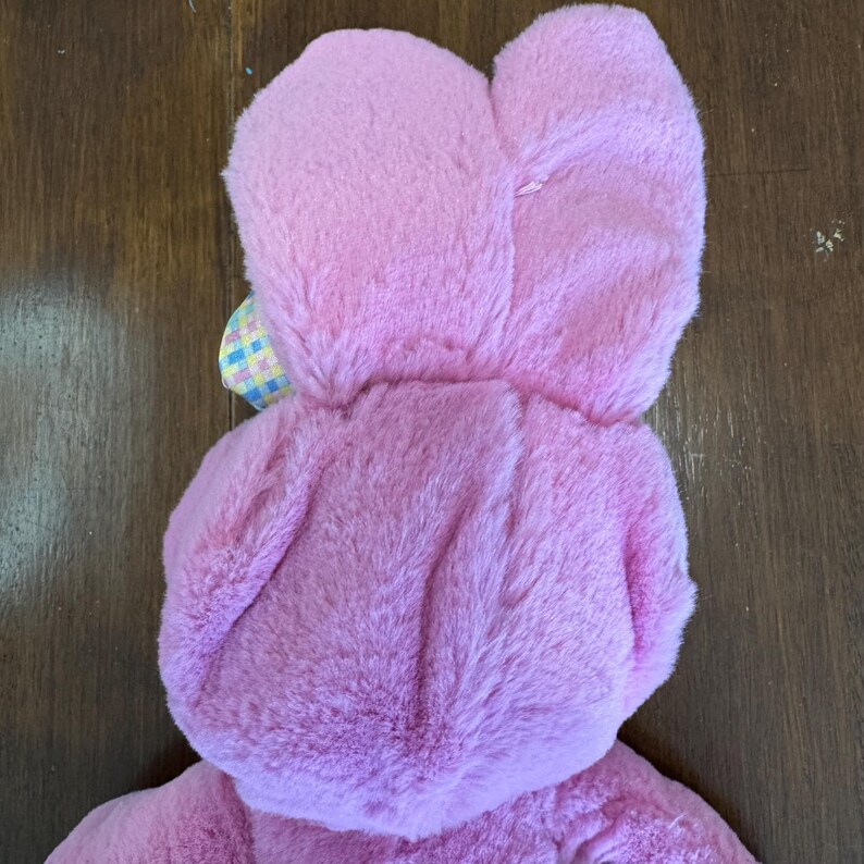 May include: A pink plush bunny costume with large ears and a hood. The costume is made of soft, fuzzy material. A small, colorful plaid bow is attached near the ears. The bunny costume is designed for children.