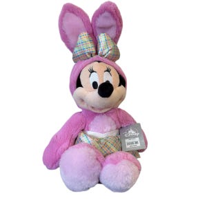 May include: A plush Minnie Mouse doll dressed in a pink bunny costume with large ears and a bow. The doll has a black nose, eyes, and a tag attached. The costume includes a hood and a patterned fabric on the belly.
