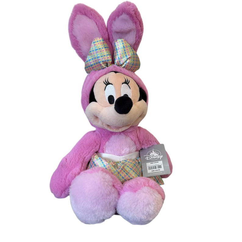May include: A plush Minnie Mouse doll dressed in a pink bunny costume with large ears and a bow. The doll has a black nose, eyes, and a tag attached. The costume includes a hood and a patterned fabric on the belly.