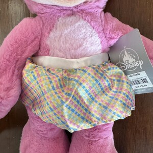 May include: A pink plush toy with a light pink belly and a pastel plaid skirt. The toy has a Disney tag attached. The skirt features a pattern of blue, yellow, and pink squares.