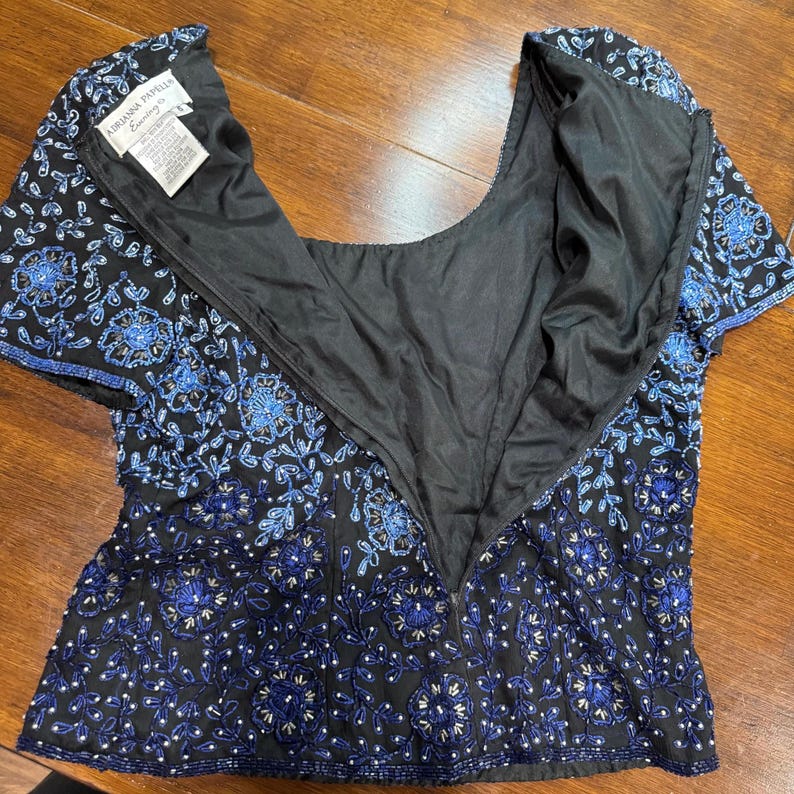 May include: Black beaded top with blue floral embroidery. The top has short sleeves and a scoop neckline. The back of the top has a zipper closure. The tag reads "Adrianna Papell Evening".