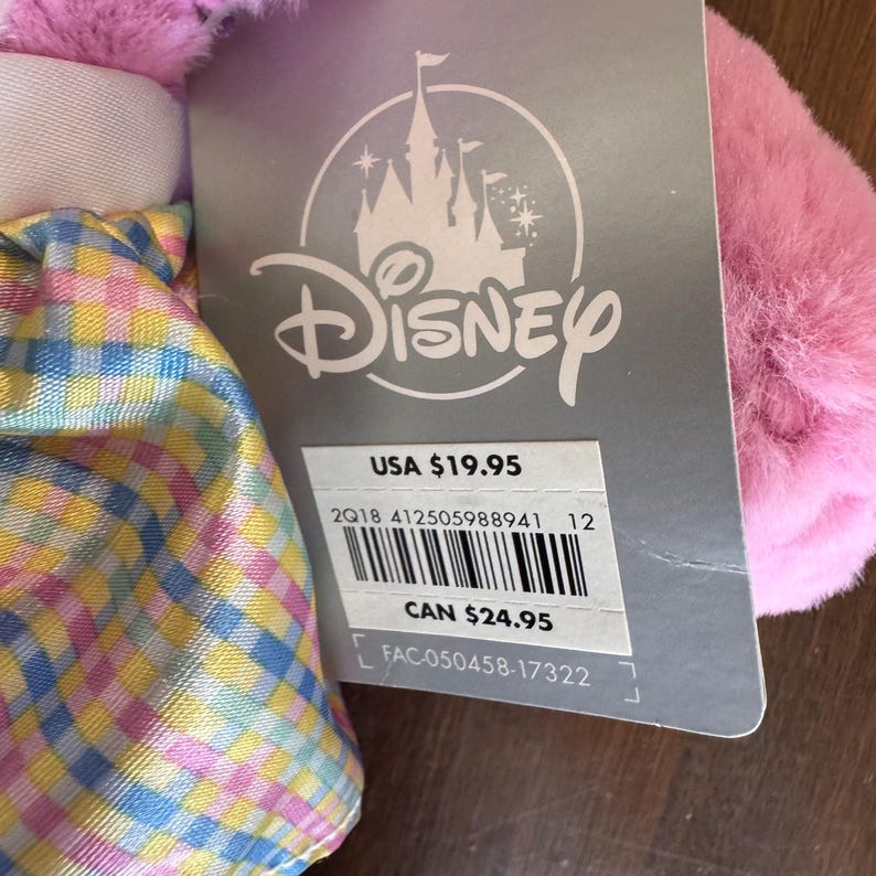 May include: A Disney product tag with the Disney logo and the text "USA $19.95". The tag is attached to a pink, fluffy item. The item has a colorful plaid pattern.