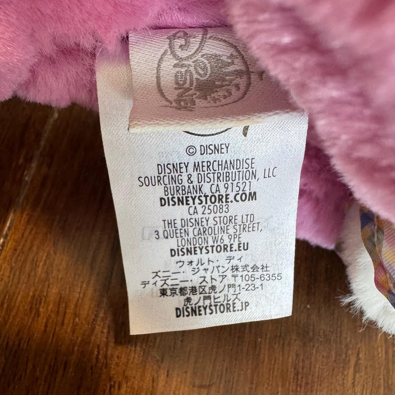 May include: A white tag with the Disney logo and text, including "DISNEY MERCHANDISE" and "DISNEYSTORE.COM". The tag is attached to a pink plush toy. The background is a wooden surface.