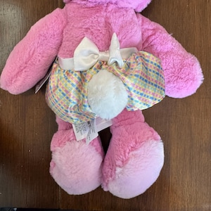 May include: A pink plush bunny toy with a white bow and a colorful plaid skirt. The bunny has a fluffy white tail and pink paws. The toy is designed for children.