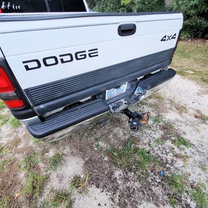 May include: White Dodge pickup truck with black lettering and a black bumper. The truck has a trailer hitch and the words "4x4" are visible. The truck is parked on a gravel and grass surface.