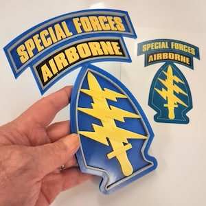 May include: A three-dimensional plastic emblem featuring the Special Forces Airborne insignia with a blue arrowhead, a yellow sword, and three yellow lightning bolts. Above it is a curved blue and yellow tab reading SPECIAL FORCES AIRBORNE.