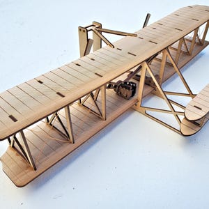 May include: A wooden model airplane with a biplane design. The model features a light brown color, with detailed construction including struts, wings, and a propeller. The model is a replica of an early aircraft.