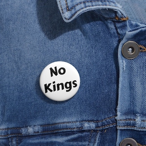 May include: A white button with the words "No Kings" in bold black letters is pinned to a blue denim jacket. The button is round and the text is centered. The jacket has visible stitching and metal buttons.