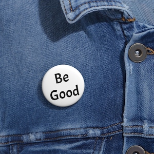 May include: A white button pin with the words "Be Good" in black, set against a blue denim jacket. The pin is round and simple, with a clear, bold font. The denim jacket has visible stitching and a button closure.