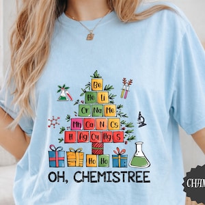 May include: A light blue t-shirt featuring a graphic of a Christmas tree constructed from colorful periodic table element squares. The design includes laboratory glassware, a microscope, and gift boxes, with the text "OH, CHEMISTREE" below.