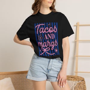May include: A black short-sleeve t-shirt featuring a graphic design with vertical blue stripes and the pink text Tacos AND margs. The design includes small pink icons of a flower, a bow, a star, and a bird, all centered on the front of the shirt.