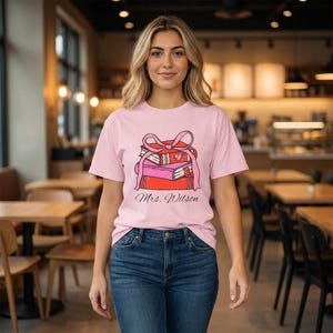 May include: A light pink t-shirt featuring a stack of books tied with a pink ribbon. The books are in shades of red and pink. The text "Mrs. Wilson" is printed below the books. The shirt is paired with blue jeans.